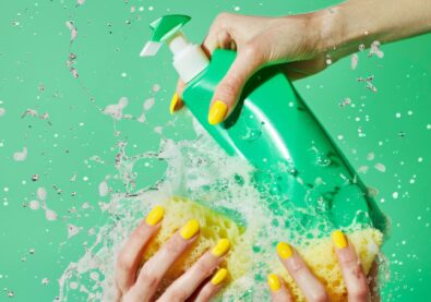 A vivid image showcasing the use of a green cleaning gel bottle; soap is being vigorously applied to a sponge. Splashes of water and soap bubbles enhance the freshness of the scene, emphasizing hygiene and cleanliness on a bright green background with a focus on detailed yellow nails.