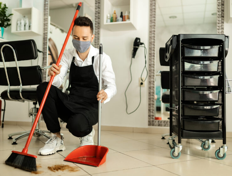 Hairstylist with protective face mask cleaning salon's floor with a broom after a haircut.