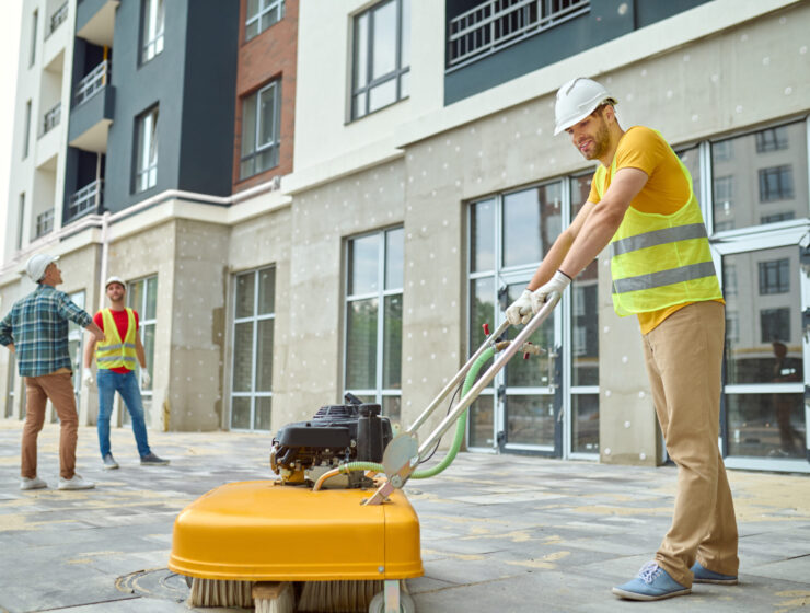 Working process. Smiling worker in protective helmet gloves and yellow tshirt cleaning construction site using special equipment and two men discussing looking at new building