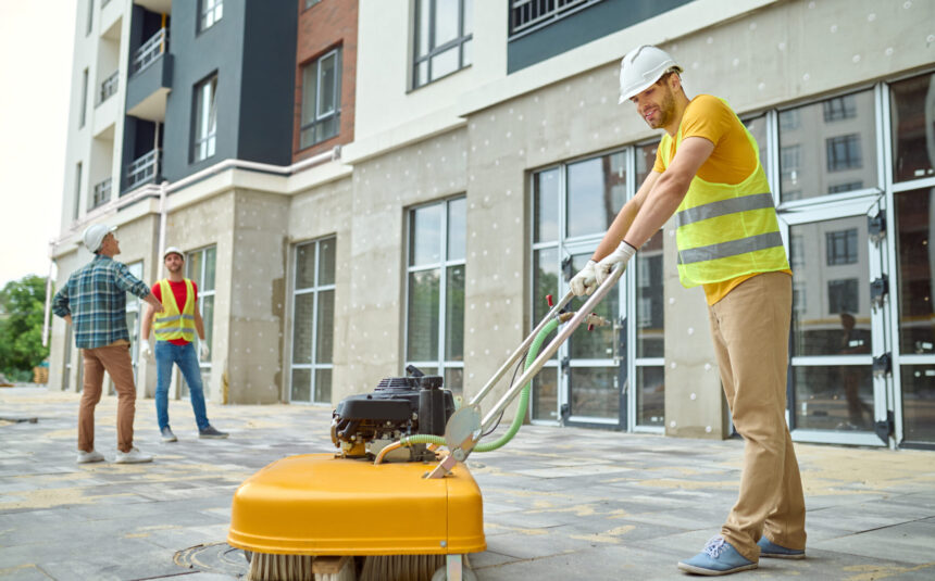 Worker cleaning construction site and two men near building Working process. Smiling worker in protective helmet gloves and yellow tshirt cleaning construction site using special equipment and two men discussing looking at new building